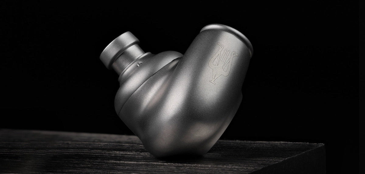 A single Meze ASTRU in-ear monitor in brushed silver, resting on a dark grained block against a near-black ground. The left third of the frame is empty black.