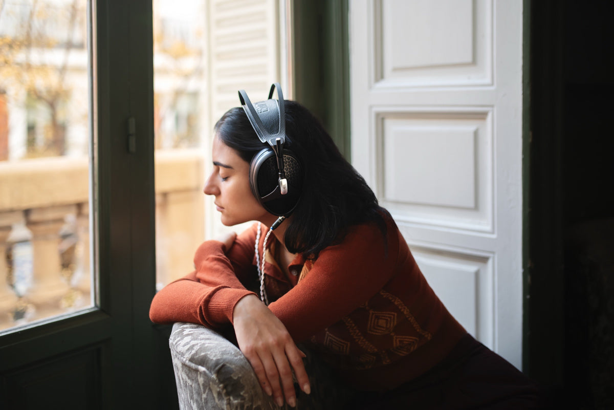 A woman in a rust orange sweater sits in a tall casement window, knees up, wearing Empyrean II, looking out at a street. Real daylight, real room, a genuine photograph rather than a studio set. Left third is the bright window, right third is a plain