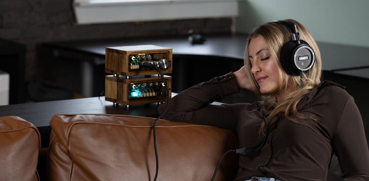 A woman on a brown leather couch listening on Grado headphones, eyes closed and one hand behind her head, two stacked wood-cased amplifier units lit up on the table behind her