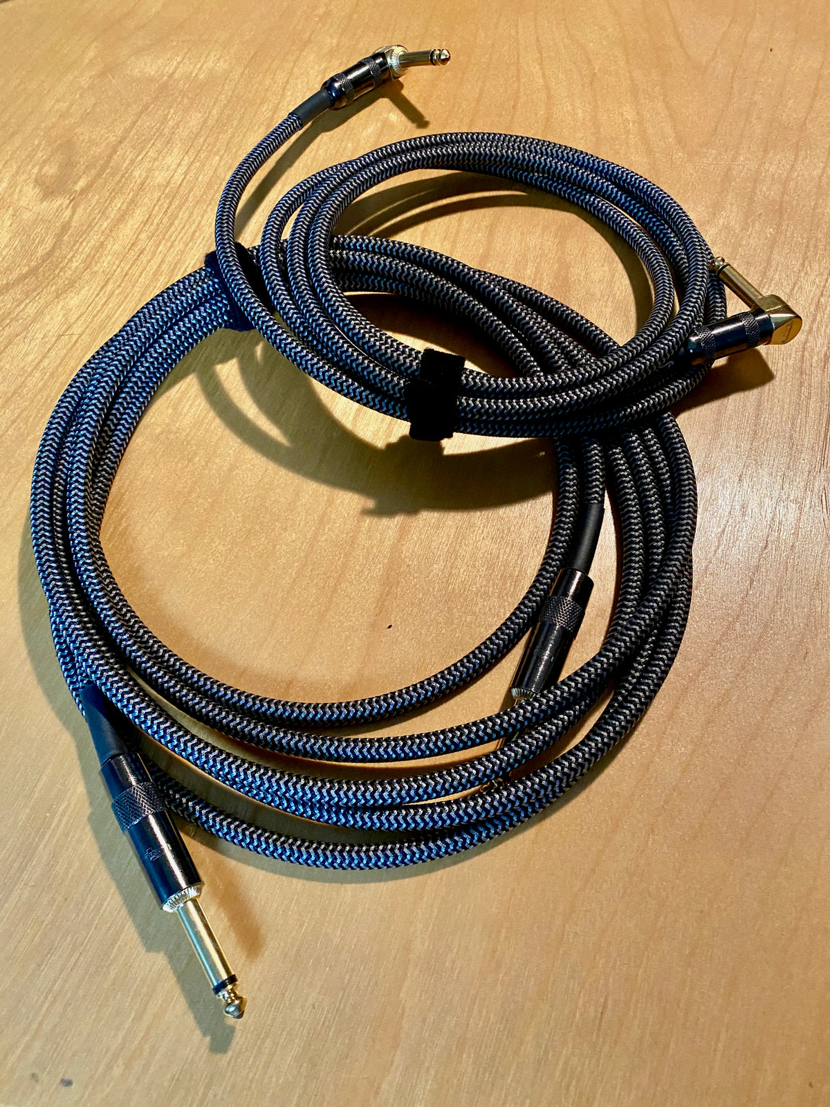 Two braided blue and black instrument cables coiled on a wooden table, each with a straight quarter-inch plug at one end and a right-angle plug at the other