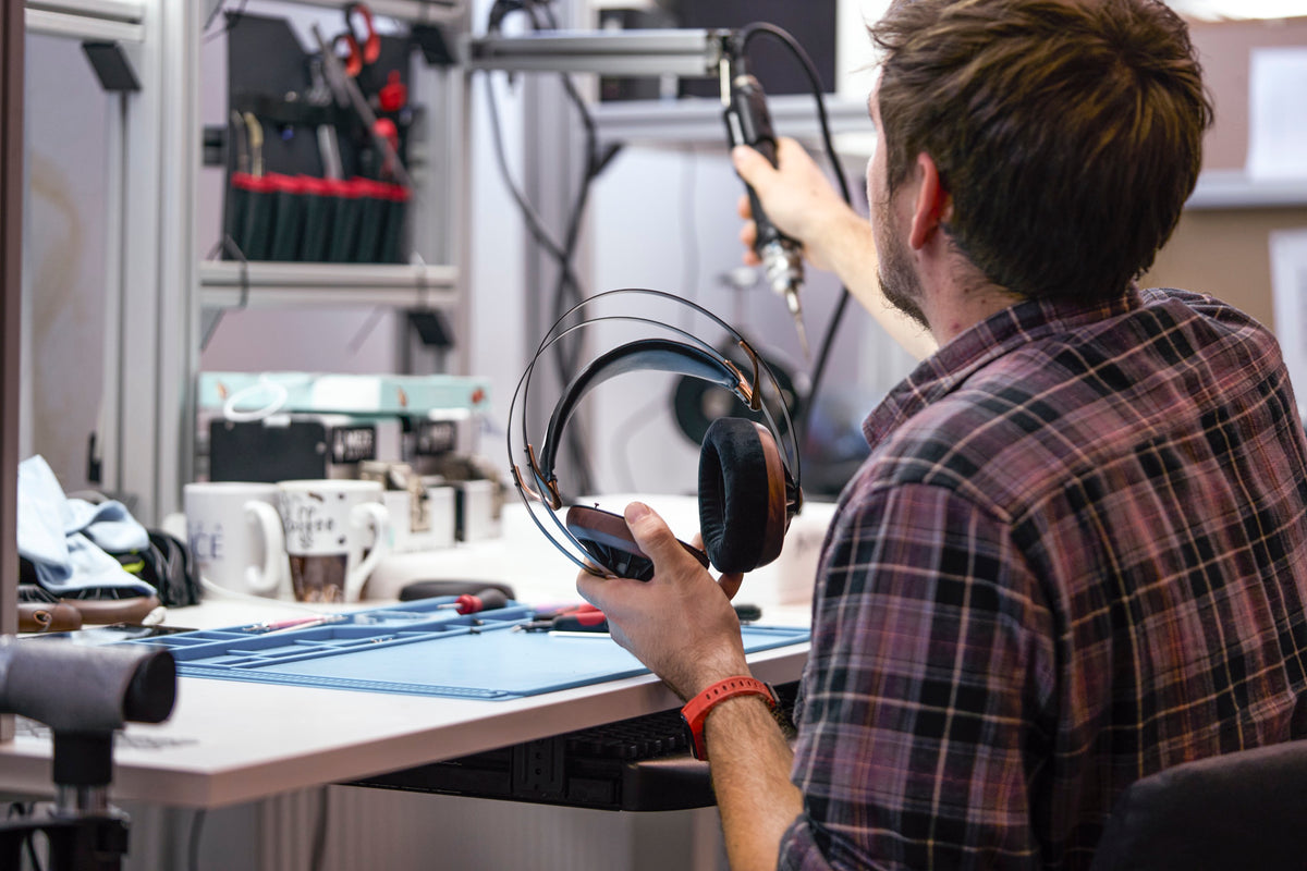 A technician in a plaid shirt at a workbench in Meze's workshop, holding a headphone in one hand and a tool in the other