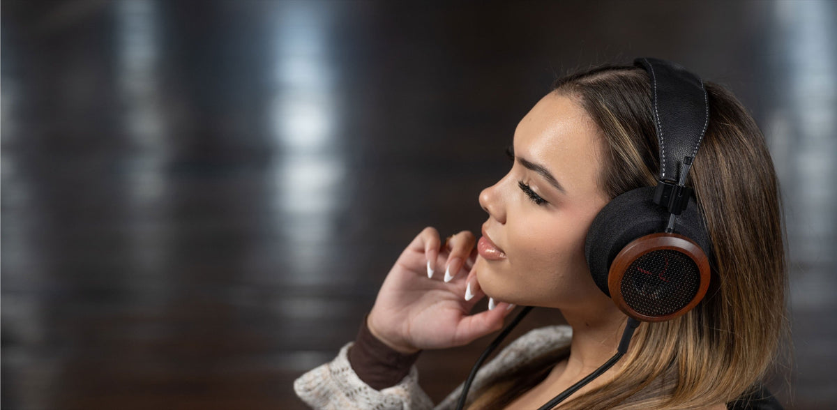 A woman in a grey knit sweater listening on wood-cup Grado headphones, eyes closed and one hand raised to the cup, in a dark room