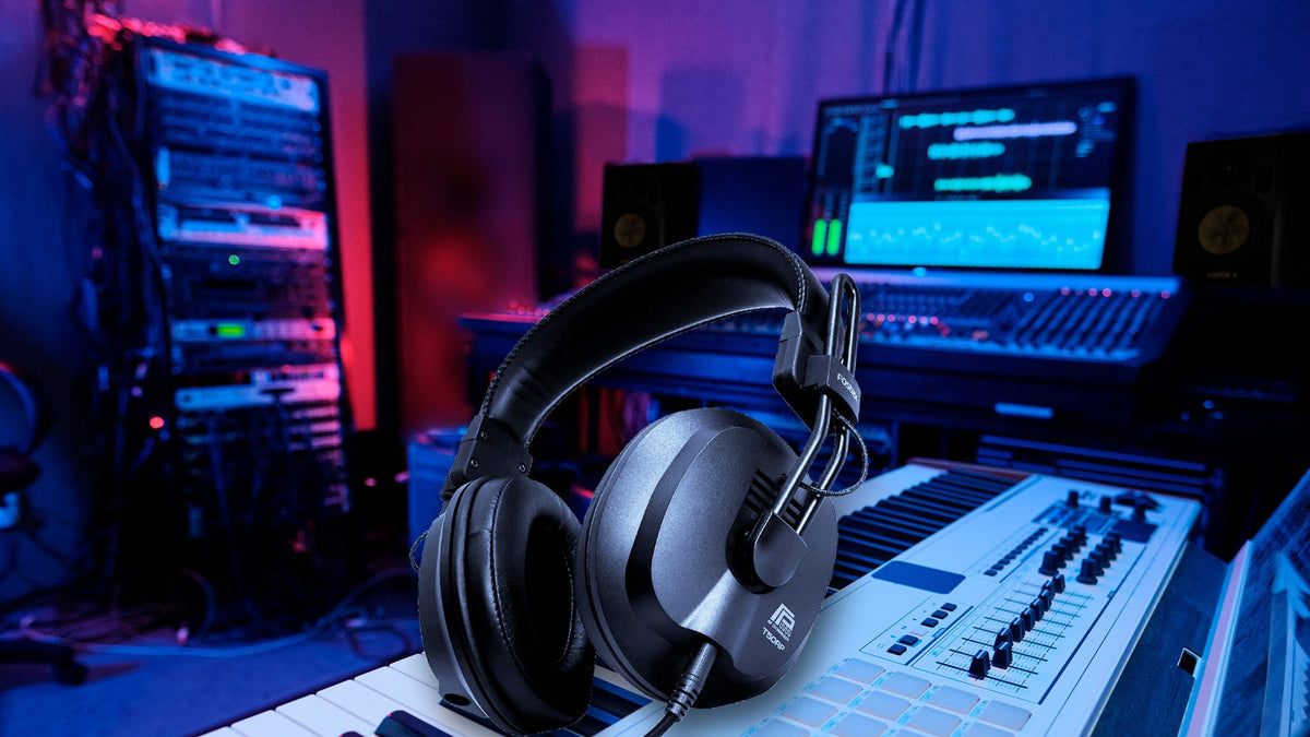 A T50RPmk4 resting on a synth keyboard in a working studio. Rack gear and a patch bay at the left, two nearfield monitors and a DAW screen behind, a control surface at the right. Real magenta and blue room light, deep shadow across the left third. Th