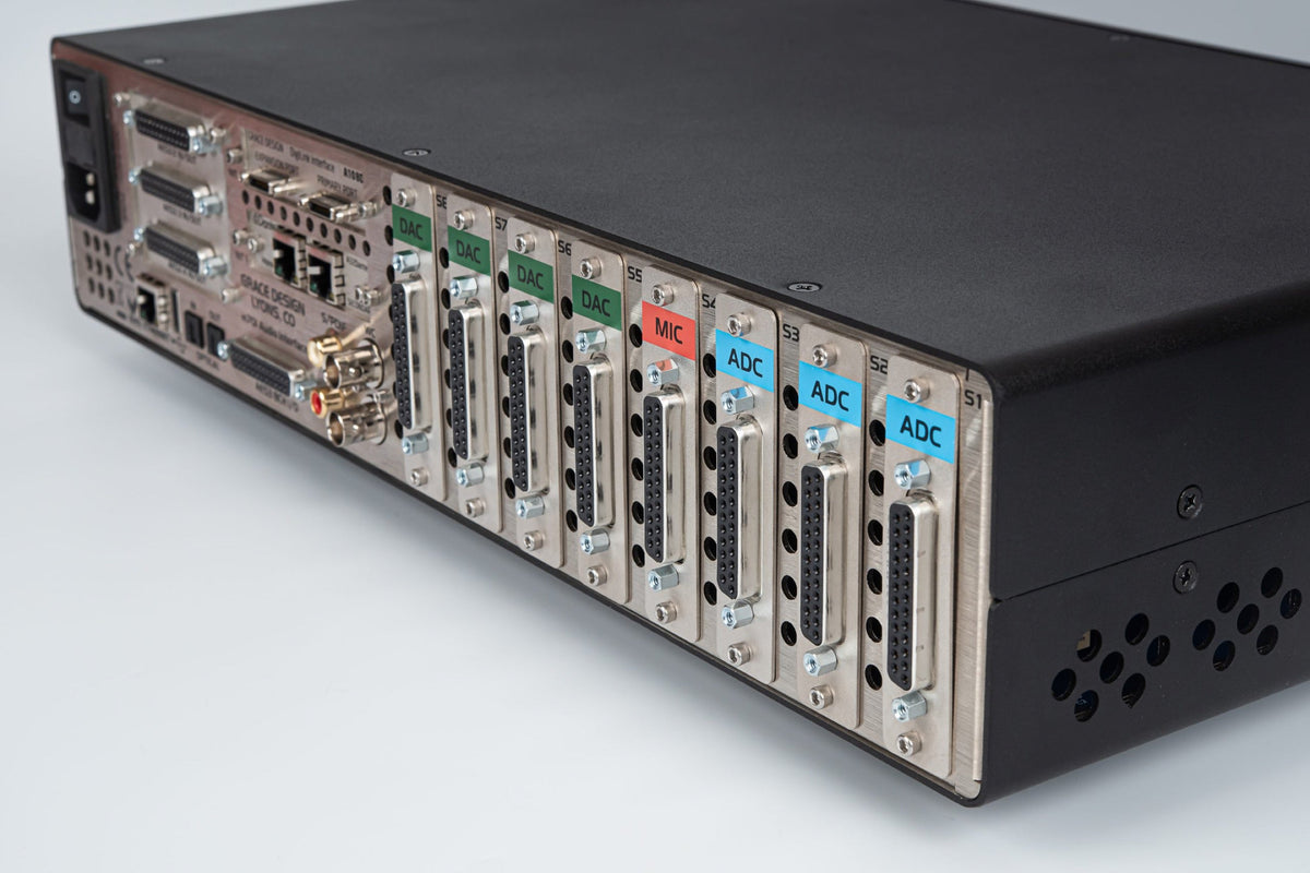 Three-quarter rear view of the m701 interface, the full width of its modular card cage filling the frame: bare aluminium module faceplates, colour-coded MIC and ADC labels, DB connectors, vent slots and the black wrap of the chassis. Cool grey ground