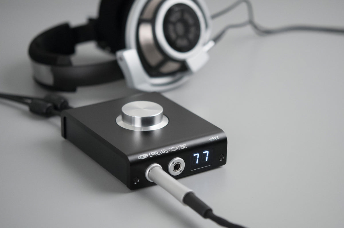 An m900 lying on a pale grey surface with its readout at 77, a light coloured headphone cable plugged in and coiling toward the lens, and a pair of silver open-back headphones (Sennheiser HD 800 by the filename) lying behind it. Low camera angle, sof