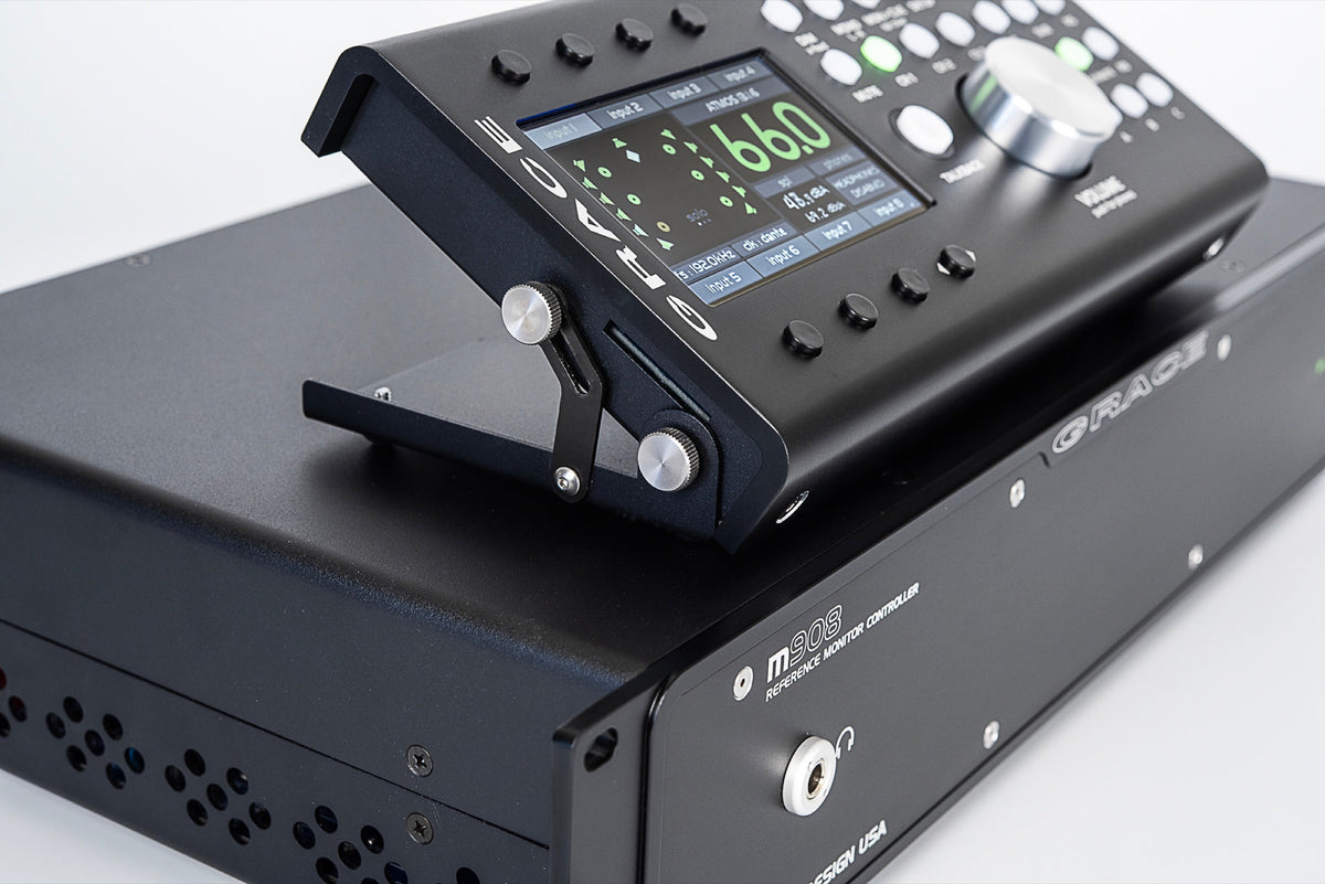 Tight three-quarter view of the m908 desktop remote tilted up on its hinged stand atop the black chassis, the colour display reading 66.0 in green, the large machined aluminium volume knob catching a highlight, and the chassis vent holes running out 