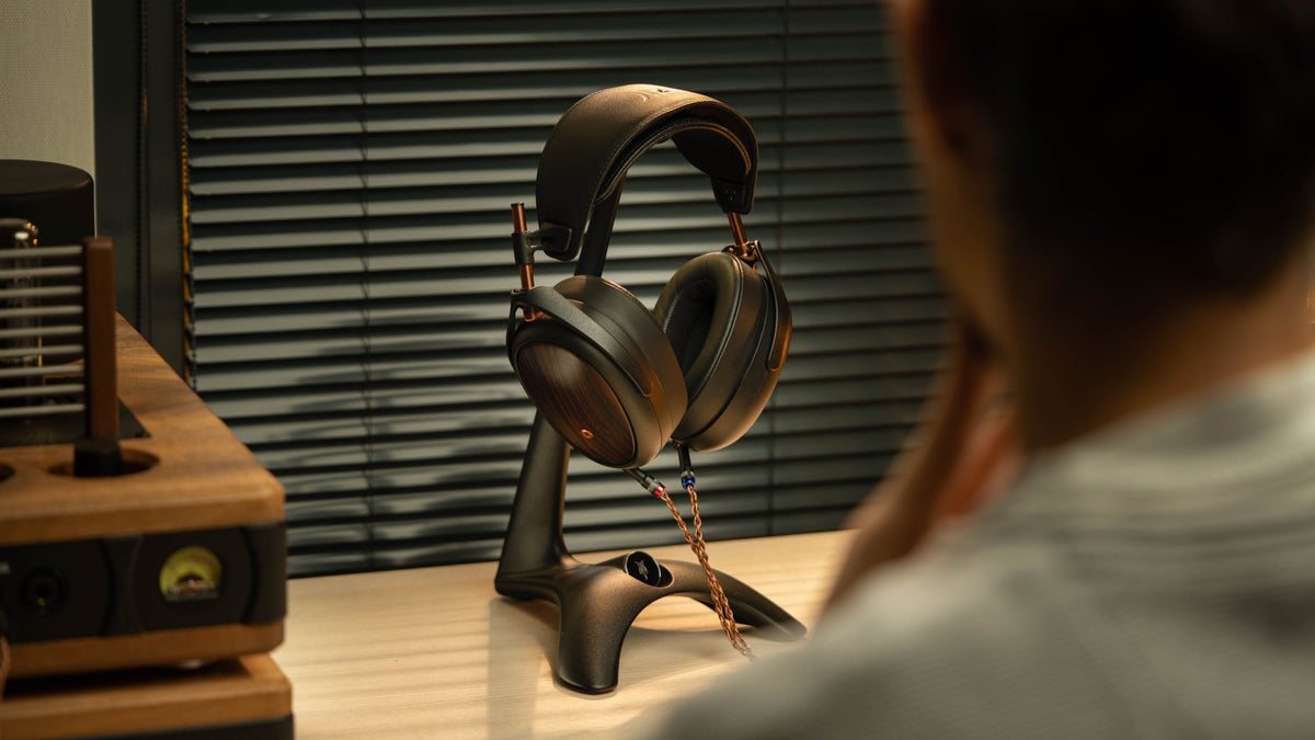 A man seen from behind at a wooden desk, headphones on a stand in front of him and a valve amplifier to the left, venetian blinds throwing warm light across the room
