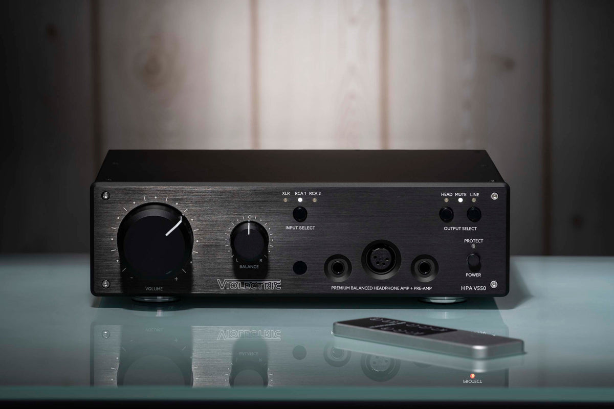 The V550 amp and its aluminium remote on a pale glass shelf with a full reflection beneath, set against a softly lit pale wood-panelled wall in a darkened room. Domestic, not studio. Generous empty space across the top third.