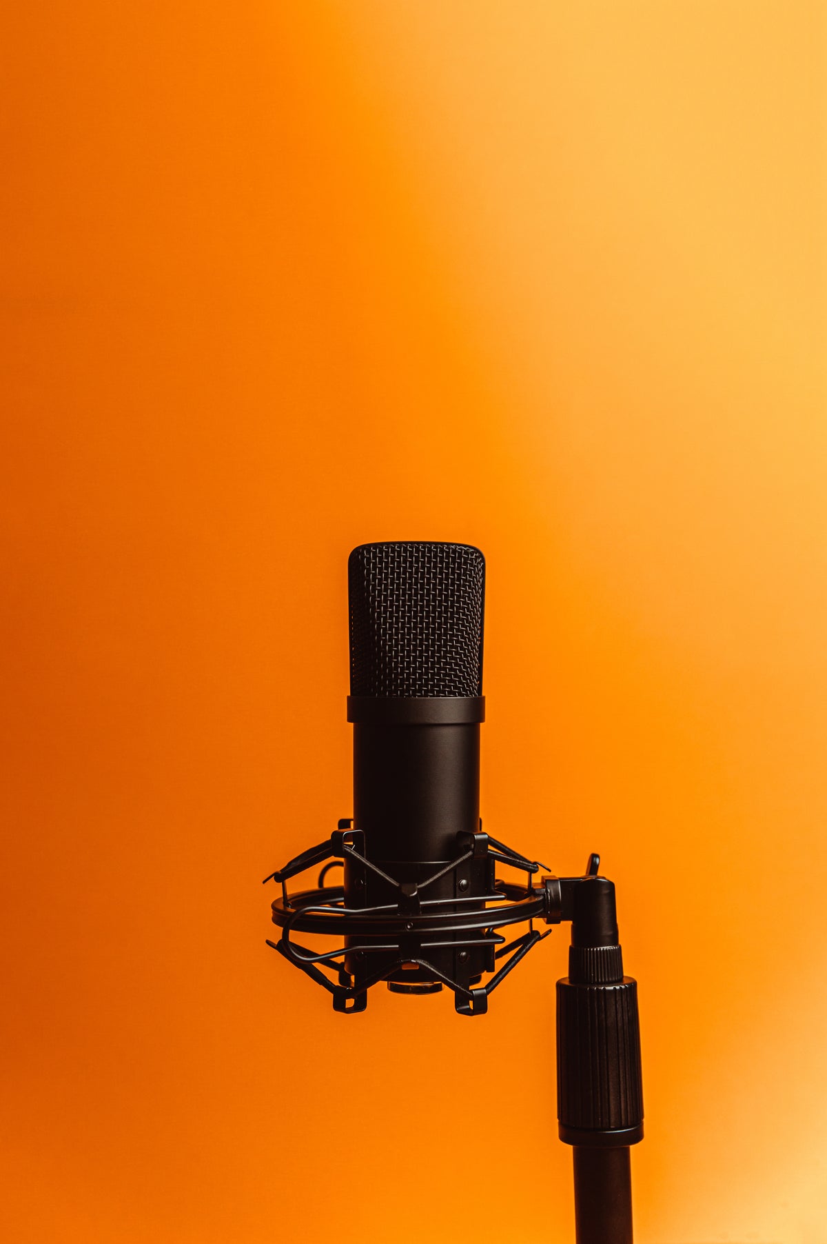 A black large-diaphragm microphone in a shock mount on a stand, against an orange wall
