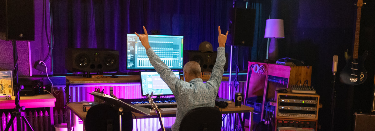 Jamey Warren at the desk at Free River Studios in Livingston, seen from behind with both hands raised, a mixer on the screen between two studio monitors and racks of outboard gear to the right