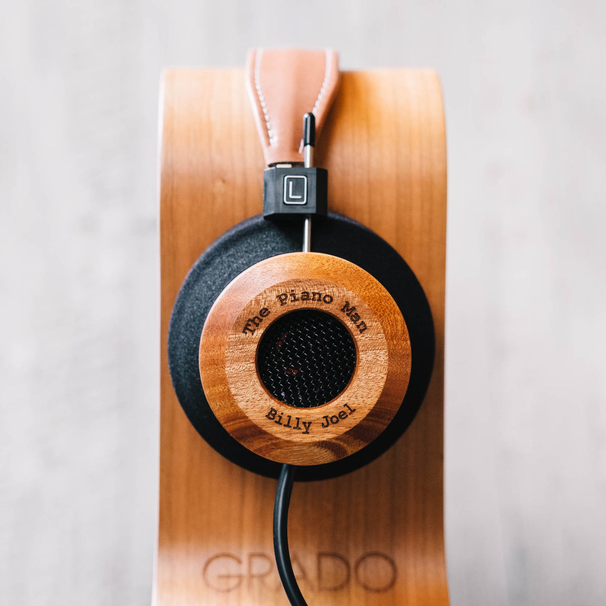 A Grado headphone with cherry wood cups and a tan leather strap hanging on a curved cherry Grado stand against a pale concrete wall