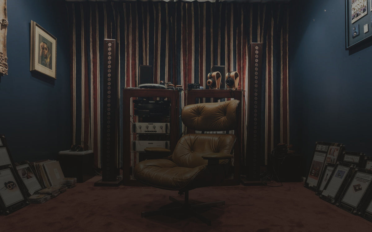 A listening room in a Grado photograph: a leather lounge chair facing the camera in front of a rack of amplifiers between two tall column speakers, two wood headphones on stands on top, and framed magazine pages leaning along the floor