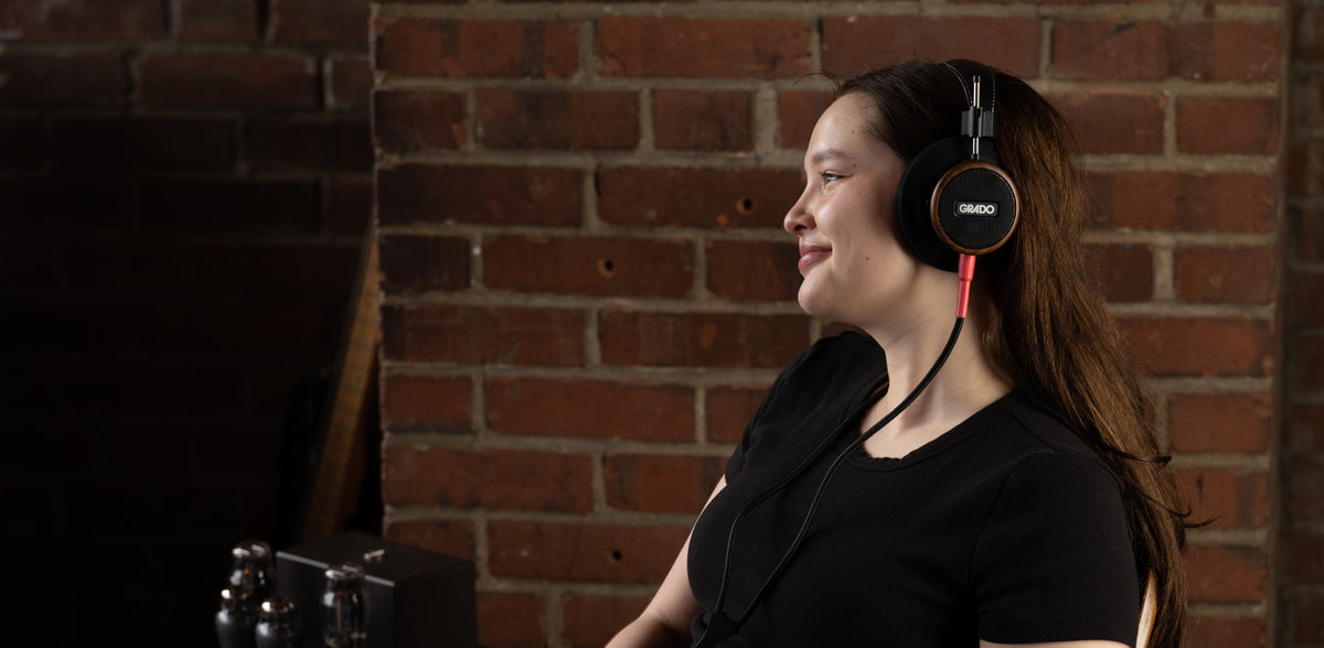 A woman in a black tee, in profile against a red brick wall, listening on Grado headphones with a red cable, a valve amplifier in the corner of the frame