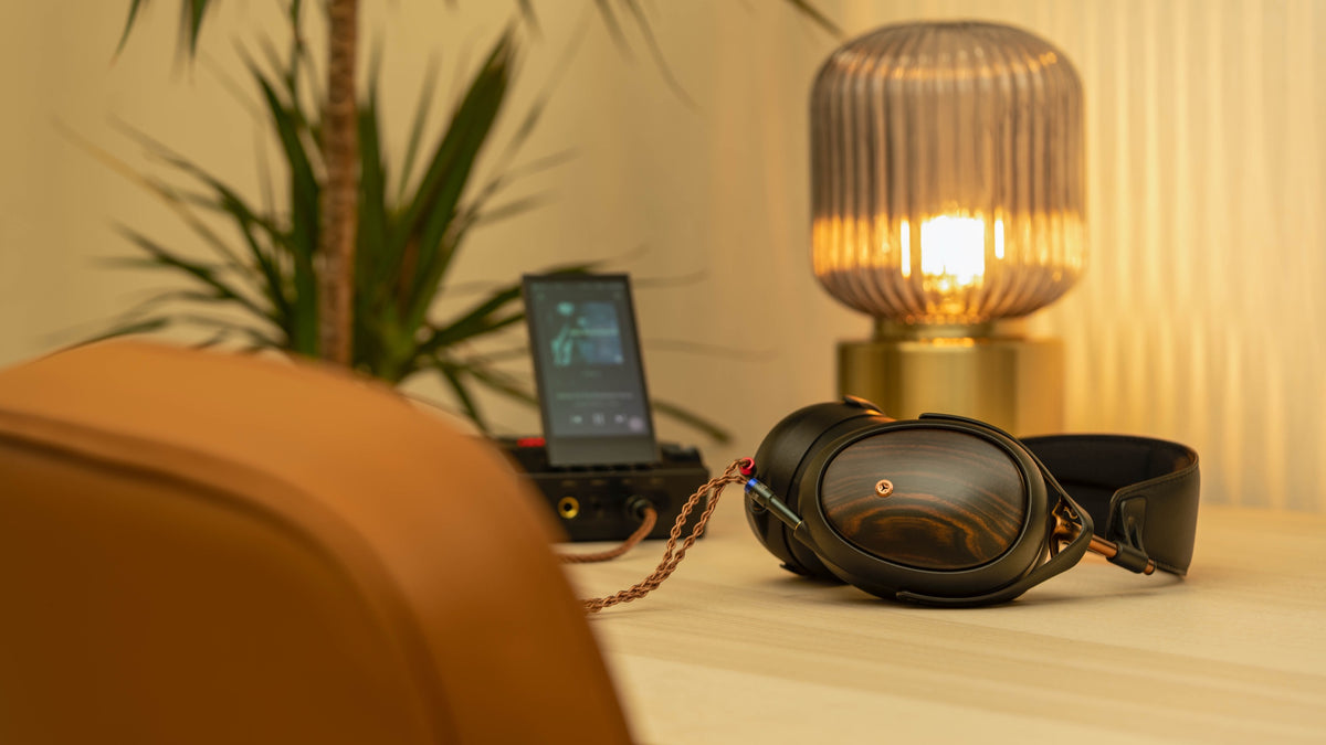 A Meze Liric lying on a desk beside a portable player, a lit fluted lamp and a plant, the back of a tan leather chair in the foreground